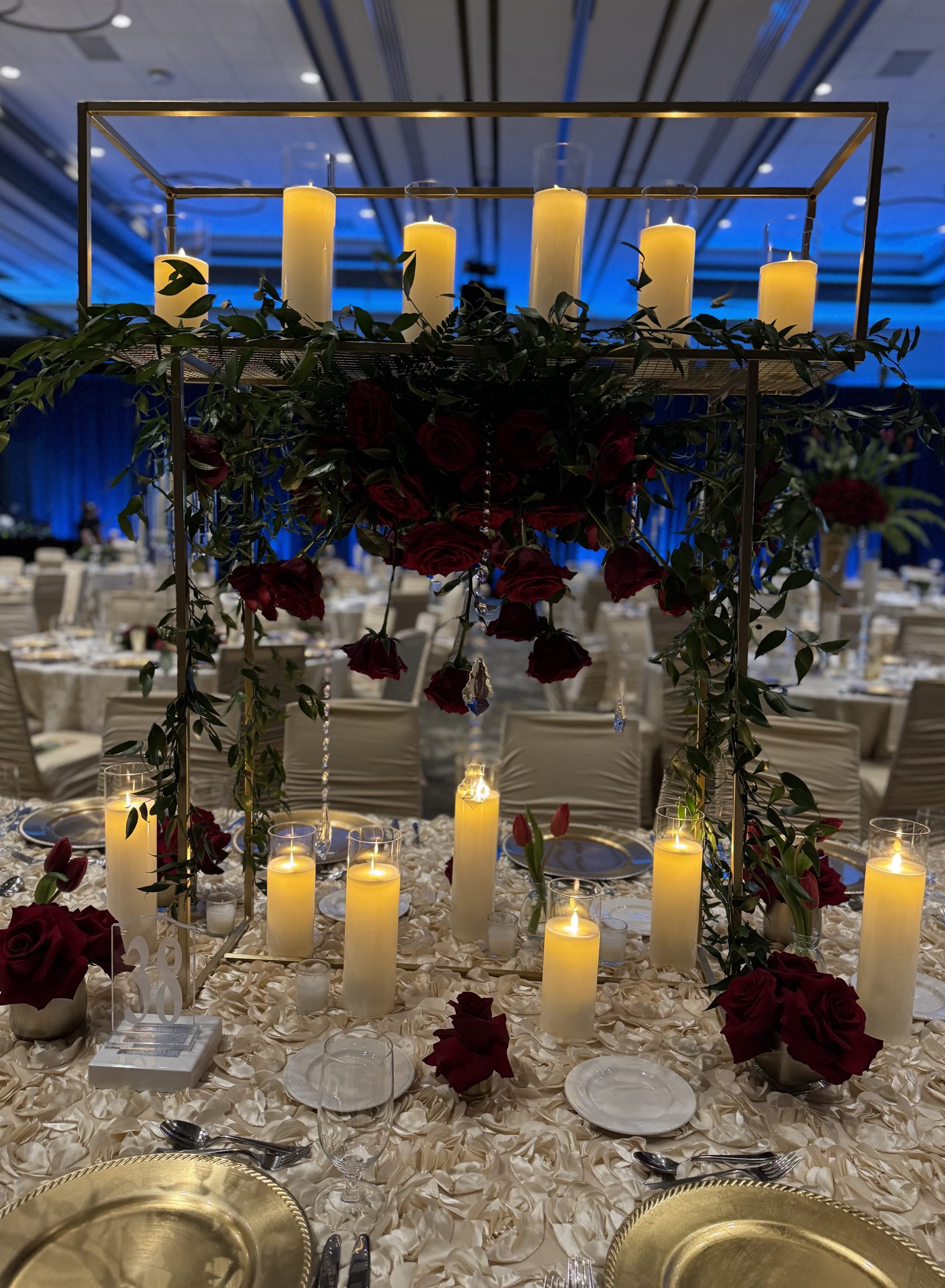 Ballroom table set with a tall gilded frame centerpiece: ivory pillar candles above a cascade of deep burgundy roses, trailing greenery and crystal strands, over an ivory rosette linen.